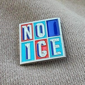 以下が含まれることがあります： 「NO ICE」の文字がグリッド状に配置された正方形のエナメルピン。文字は白で、赤と青のアクセントが付いています。背景は、質感のあるニュートラルな色合いの生地です。