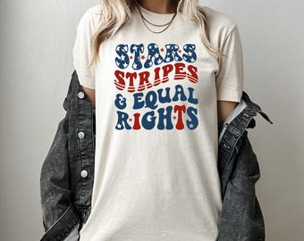 May include: Cream-colored t-shirt with the text "STARS STRIPES & EQUAL RIGHTS" in blue and red. The word "STARS" has blue stars as the letter fill. The shirt is worn with a black denim jacket.