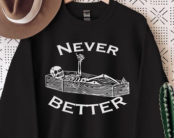 May include: Black sweatshirt with the words "NEVER BETTER" in white, featuring a skeleton in a coffin. A brown hat hangs to the left, and a cactus in a white pot sits on a wooden table to the right.