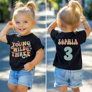 Può includere: Una maglietta nera con la scritta "YOUNG WILD & THREE" in un font retrò colorato. La parte posteriore della maglietta ha il nome "SOPHIA" e il numero "3". Il bambino indossa pantaloncini di jeans.