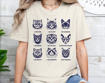 May include: Beige t-shirt featuring nine cat faces with various expressions and accompanying text. Phrases include "I love you," "I am happy," and "Just curious." The design is in dark blue.