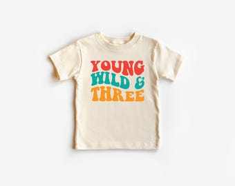May include: Cream-colored toddler t-shirt with the words "YOUNG WILD & THREE" in red, teal, and orange retro font. The short-sleeved shirt is centered on a white background.