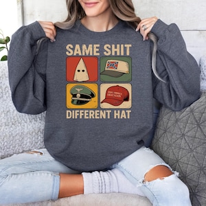 May include: Dark grey sweatshirt featuring the text "SAME SHIT DIFFERENT HAT" and four hat illustrations. The images include a white hood, a Confederate flag hat, a military hat, and a red "Make America Great Again" hat.