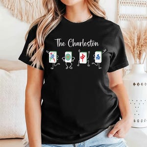 May include: Black t-shirt with the text "The Charleston" above a graphic of four animated Mahjong tiles. The tiles are white with colorful designs and have stick figure arms and legs.