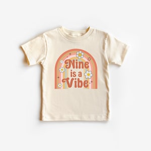 Puede incluir: Camiseta color crema con un diseño de arcoíris retro. El gráfico incluye el texto "Nine is a Vibe" en una fuente lúdica, junto con margaritas y estrellas. Una camiseta divertida y colorida para niños.