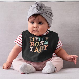 May include: A baby wearing a black bib with the words "LITTLE BOSS LADY" in colorful retro font. The baby is also wearing a gray turban, a striped shirt, pink pants, and gray socks. The baby is sitting on a gray couch.