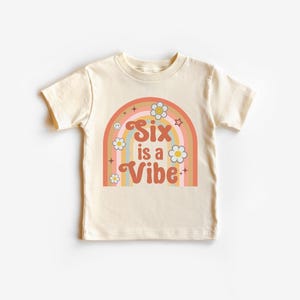 以下が含まれることがあります： レトロな虹のデザインが施された、クリーム色の半袖Tシャツ。「Six is a Vibe」の文字がグルーヴィーなフォントで、花と星のグラフィックとともに描かれています。子供向けの楽しくてカラフルなTシャツです。