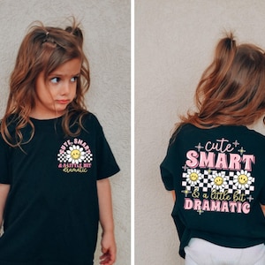 May include: Black t-shirt featuring the text "cute, smart & a little bit dramatic" and a daisy graphic. The shirt is displayed on a young girl. The back of the shirt also has the same text and daisy graphic.