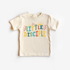 Könnte beinhalten: Ein cremefarbenes T-Shirt mit dem Schriftzug "LITTLE DISCIPLE" in bunten, verspielten Buchstaben. Das Design umfasst ein Kreuz, eine Sonne, Blitze und eine Krone.