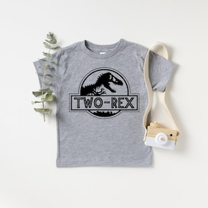以下が含まれることがあります： 黒い恐竜のグラフィックと「TWO-REX」の文字が入ったグレーの半袖Tシャツ。 デザインは円と長方形のボックスに入っています。 このTシャツは2歳児向けにデザインされています。