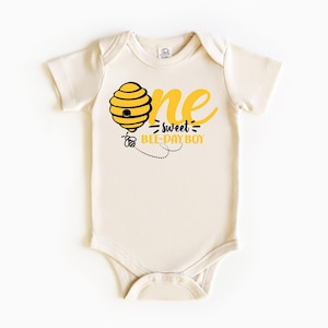May include: Cream-colored baby bodysuit with a yellow and black bee-themed graphic. The design includes a beehive, a bee, and the text "one sweet bee-day boy" in yellow and black. The bodysuit has short sleeves and a snap closure.