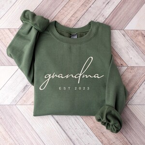 Pode incluir: Um moletom verde com a palavra "grandma" em fonte cursiva. Abaixo, o texto diz "EST 2023". O moletom está sobre uma superfície de madeira clara com um padrão de espinha de peixe.