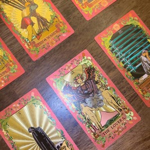 May include: A collection of tarot cards with pink borders and gold accents. The cards feature detailed illustrations and text, including "Ten of Wands," "Queen of Cups," and "The Lovers." The cards are arranged on a wooden surface.
