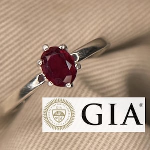 May include: A silver ring featuring a vibrant, oval-cut ruby gemstone. The ruby is set in a classic prong setting, and the ring band is a simple, polished silver. The GIA logo is visible in the image.
