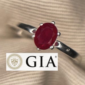 以下が含まれることがあります： 鮮やかなオーバルカットのルビーの宝石をあしらったシルバーリング。 ルビーは4本爪でセットされ、リングバンドは滑らかで磨かれています。 GIAのロゴが画像に表示されています。