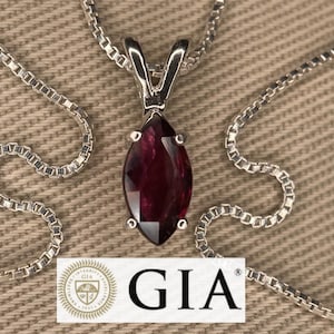 Puede incluir: Un collar de plata con un colgante de piedra preciosa roja oscura con forma de marquesa. El colgante cuelga de una delicada cadena. El logotipo y el texto de GIA son visibles en la parte inferior de la imagen.