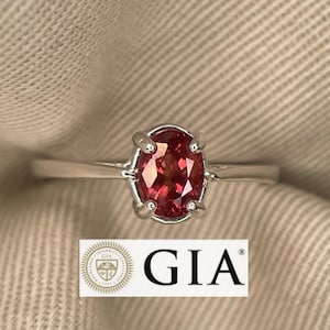 Pode incluir: Um anel de prata com uma pedra preciosa ovalada rosa-avermelhada. O anel tem quatro garras e está sobre um tecido de cor neutra. O logotipo e o texto GIA são visíveis abaixo.