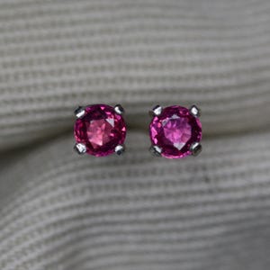 May include: A pair of round, faceted, deep pink gemstone stud earrings set in silver-toned metal prongs. Each stone features a brilliant cut that reflects light, showcasing a vibrant magenta hue. The earrings are positioned side by side.