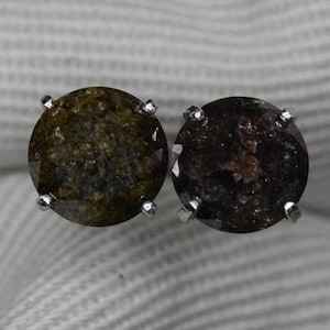 May include: A pair of round stud earrings. Each earring features a dark, speckled gemstone set in a silver metal prong setting. The gemstones have a mix of olive green and dark brown tones.