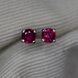 May include: A pair of round, faceted, deep red ruby stud earrings set in four-prong silver metal settings. Each gemstone features a brilliant cut that reflects light, showcasing a vibrant crimson hue and a polished, metallic finish on the prongs.
