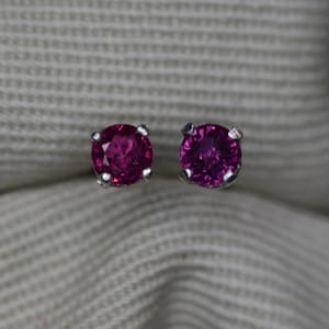 May include: A pair of stud earrings featuring round, faceted, deep pink-red gemstones set in silver-toned metal prongs. Each earring has a classic four-prong setting that secures the circular stone, highlighting the vibrant color and reflective facets.