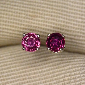 May include: A pair of stud earrings featuring round, vibrant pink gemstones set in silver-colored metal. The earrings are displayed on a beige, textured fabric, showcasing their color and design.