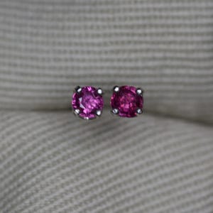 May include: A pair of round, faceted, deep pink gemstone stud earrings set in four-prong silver metal settings. The stones are circular in shape with a brilliant cut, reflecting light from their polished surfaces, and are mounted on silver posts.