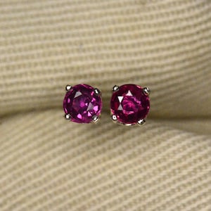 May include: A pair of stud earrings featuring round, faceted, deep pink gemstones set in silver. The earrings are displayed on a beige, textured fabric background.