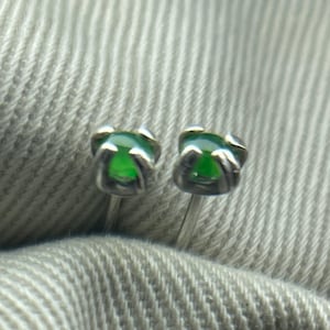 May include: A pair of small stud earrings featuring bright green gemstones set in silver-coloured metal. The gemstones are rounded and held in place by a four-prong setting. The earrings are shown on a textured, light grey fabric.