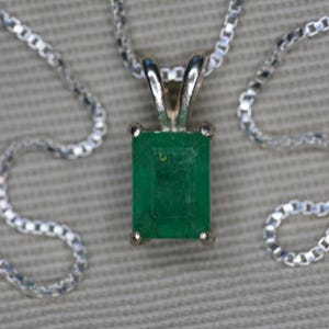 May include: A silver necklace with a rectangular emerald pendant. The emerald is a vibrant green color and is set in a silver bezel. The chain is a delicate box chain style.