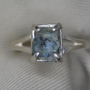Puede incluir: Un anillo de plata con una piedra preciosa rectangular de color azul claro. La piedra preciosa está engastada en un engaste de cuatro puntas, y la banda se divide en dos a cada lado de la piedra.