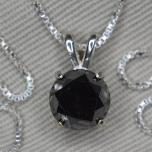Puede incluir: Un collar de cadena de plata con un colgante de diamante negro. El diamante es redondo y tiene un corte facetado.