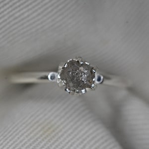 May include: A silver ring with a rough, gray diamond set in a prong setting.