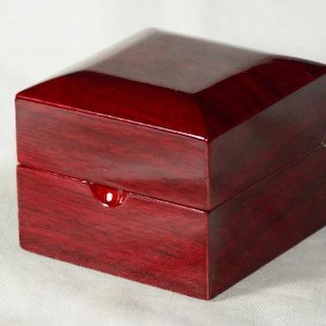 May include: A square, wooden box with a glossy, dark red finish. The box has a hinged lid and a small, rounded indentation on the front. The box is likely used for jewelry or other small items.