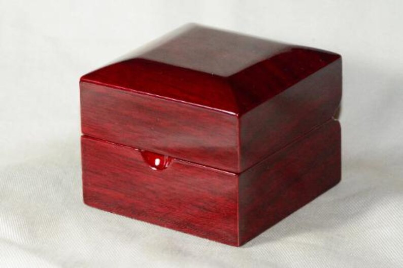 May include: A square, wooden box with a glossy, dark red finish. The box has a hinged lid and a small, rounded indentation on the front. The box is likely used for jewelry or other small items.