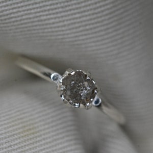 May include: A silver ring with a rough, gray diamond set in a prong setting.
