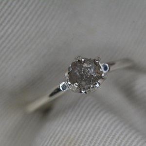 May include: A silver ring with a rough, gray diamond set in a prong setting.