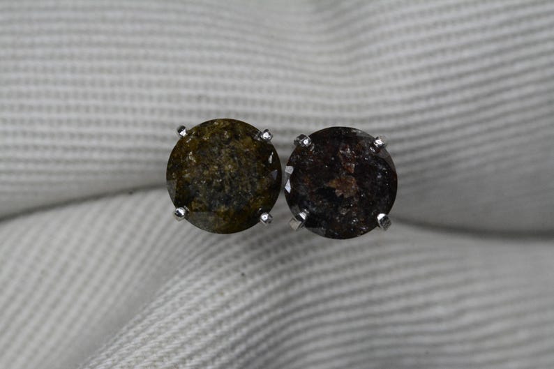 May include: A pair of round stud earrings. Each earring features a dark, speckled gemstone set in a silver metal prong setting. The gemstones have a mix of olive green and dark brown tones.