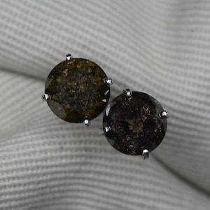 May include: A pair of round stud earrings with dark, speckled gemstones. Each earring features a silver-colored setting with four prongs holding the stone. The gemstones have a mix of dark green and brown hues.