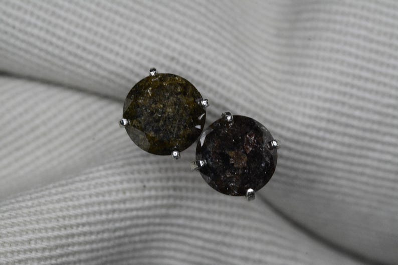 May include: A pair of round stud earrings with dark, speckled gemstones. Each earring features a silver-colored setting with four prongs holding the stone. The gemstones have a mix of dark green and brown hues.