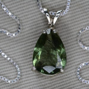 May include: A silver necklace featuring a pear-shaped, green gemstone pendant. The pendant is faceted and has a dark, internal pattern. The chain is a delicate, linked design. The necklace is displayed on a textured, light gray surface.