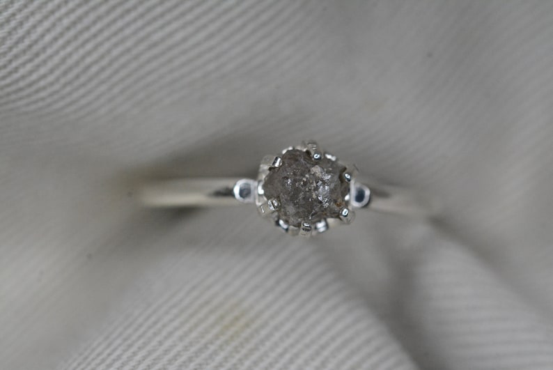 May include: A silver ring with a rough, gray diamond set in a prong setting.
