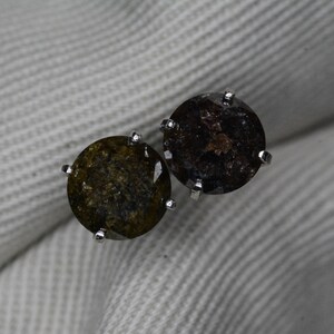 May include: A pair of round stud earrings. Each earring features a dark, speckled gemstone set in a silver-colored prong setting. The gemstones have a mix of dark brown, olive green, and black tones. The earrings are displayed on a white, textured surface.