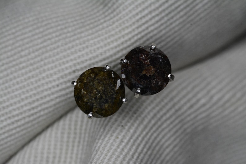 May include: A pair of round stud earrings. Each earring features a dark, speckled gemstone set in a silver-colored prong setting. The gemstones have a mix of dark brown, olive green, and black tones. The earrings are displayed on a white, textured surface.