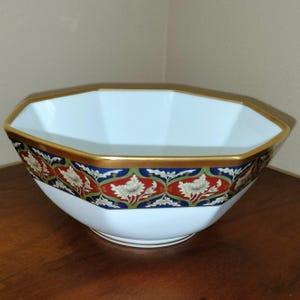 May include: An octagonal white porcelain bowl featuring a gold-trimmed rim and a decorative band around the exterior. The band displays a repeating pattern of stylized white floral motifs set against alternating red and blue panels with green accents.