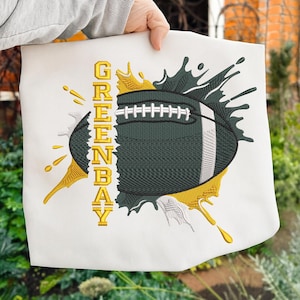 Könnte beinhalten: Weißer Stoff mit einem gestickten Football-Design. Der Football ist dunkelgrün mit weißen Schnürsenkeln, und der Text "GREENBAY" ist in Gold gehalten. Goldene und dunkelgrüne Farbspritzer umgeben den Football.