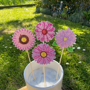 May include: Four decorative flower ornaments with pink and purple beaded petals and yellow centres are displayed in a white pot. The flowers are on wooden sticks, set against a backdrop of green grass and sunlight.