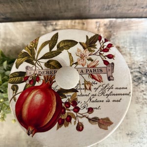 May include: A round, off-white decorative lid featuring a large red pomegranate and floral accents. The lid has a wooden knob and text that reads "DEBER A PARIS" and a quote about life and nature.