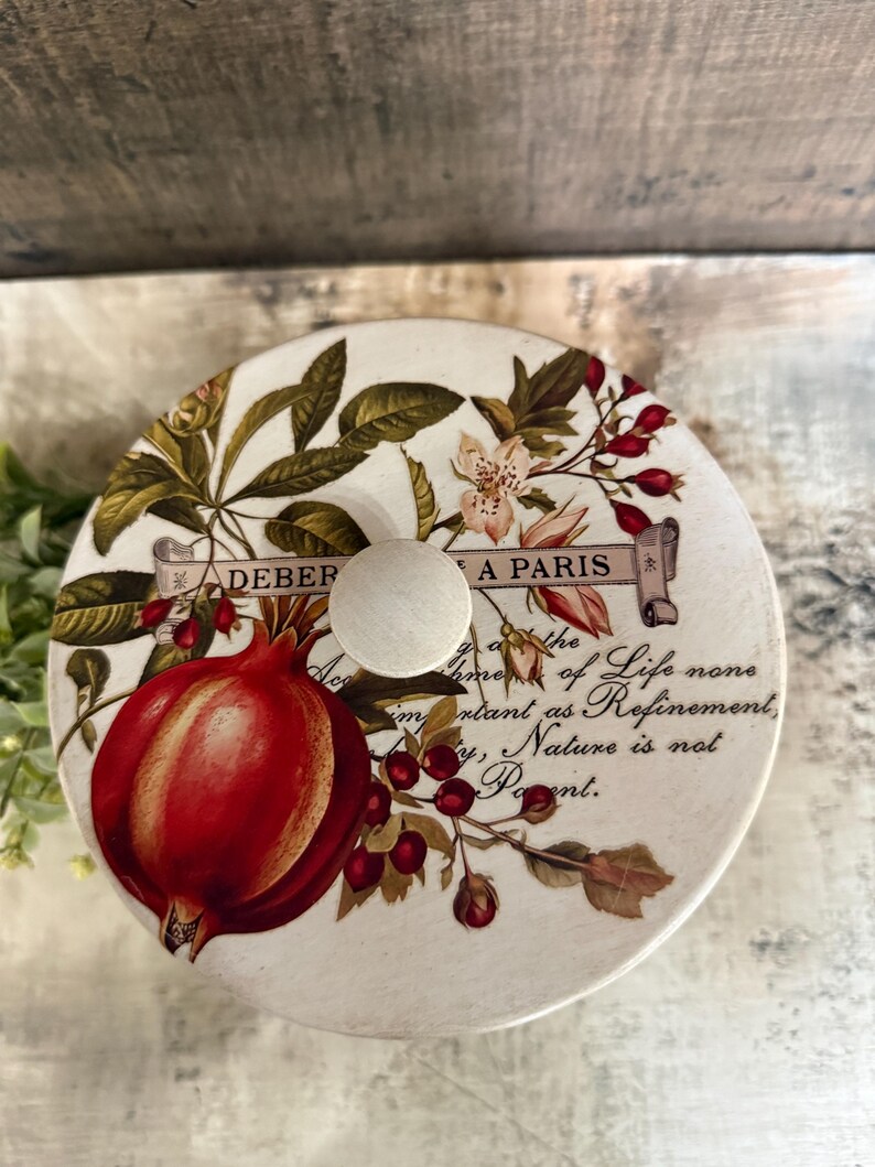 May include: A round, off-white decorative lid featuring a large red pomegranate and floral accents. The lid has a wooden knob and text that reads "DEBER A PARIS" and a quote about life and nature.