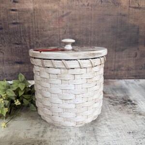May include: A cream-colored woven basket with a lid. The lid has a small, round knob and a painted design. The basket is cylindrical and has a textured, woven appearance. The background is a rustic wooden surface.